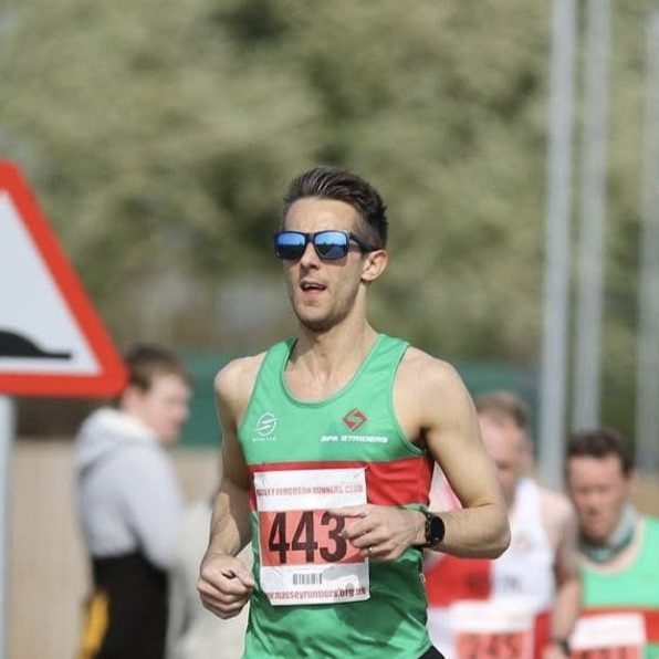 ImgDR20230923v1 cropped square White male with brown hair and sunglasses on, running in a green vest with race number and black shorts