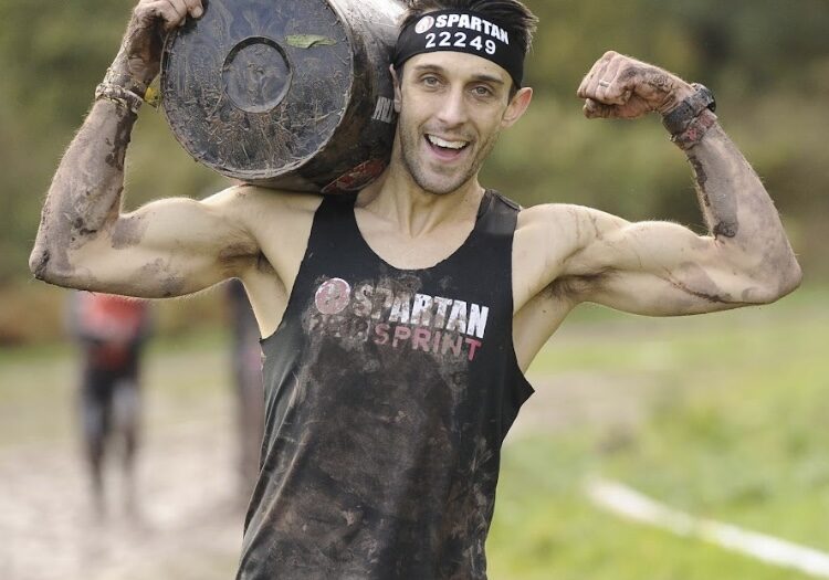White male with brown hair, smiling flexing biceps while carrying a barrel and covered in mud